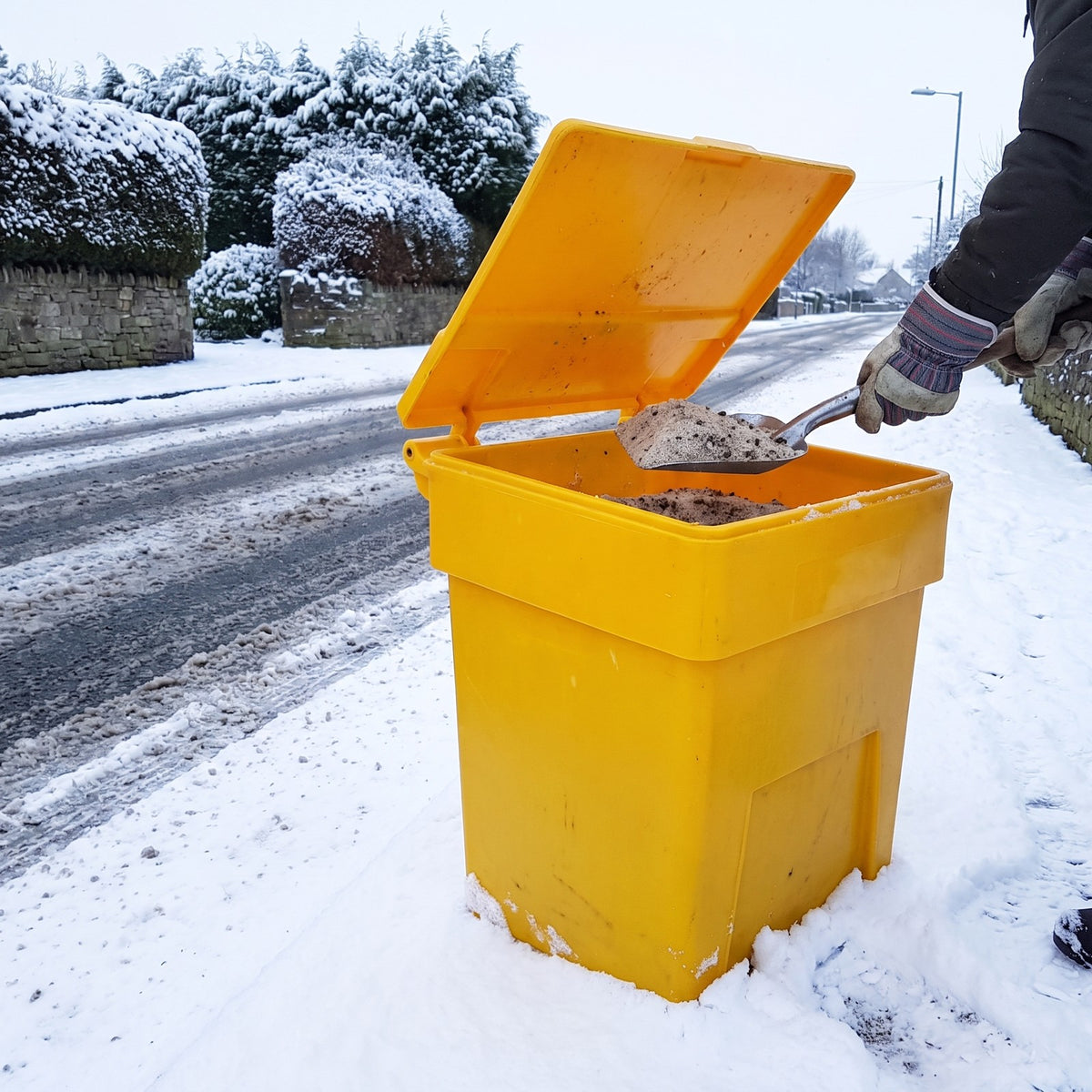 Grit being scooped from a yellow 200L grit bin at a snowy roadside walkway
